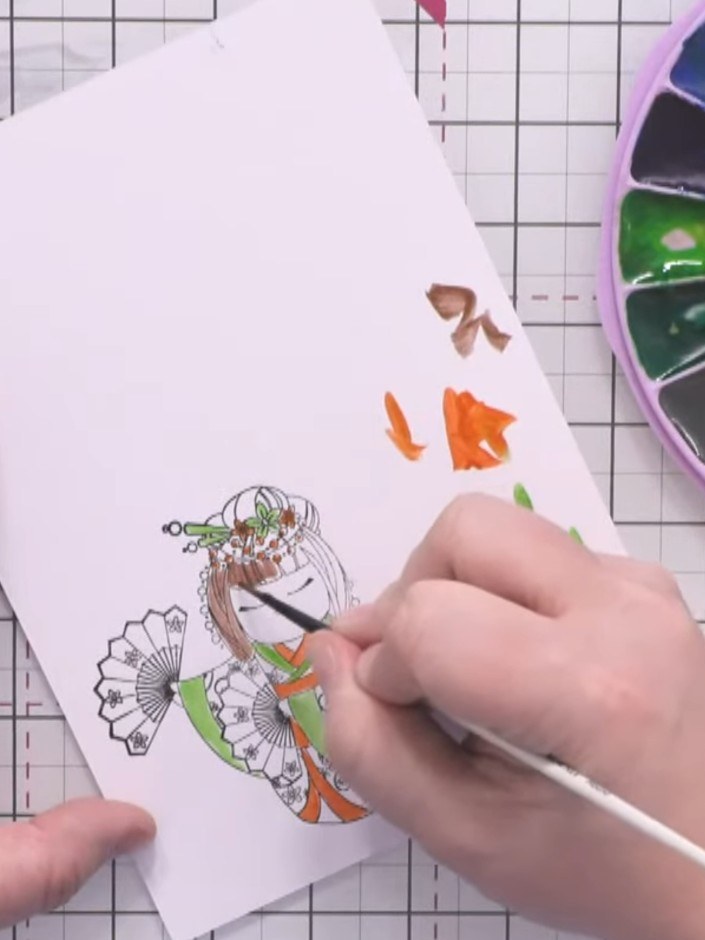 Step 9: Paint the character's hair in brown using watercolour. Add facial details with the brush. Colour the fan in gr