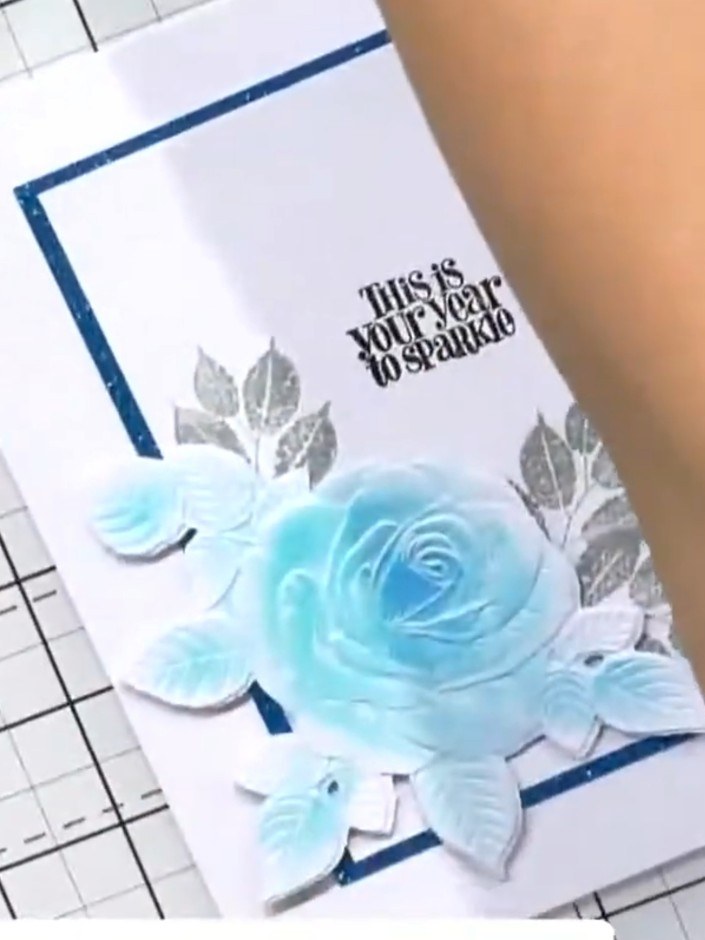 Step 5: Position and adhere a white embossed flower element to the top corner of the card, overlapping the blue frame 