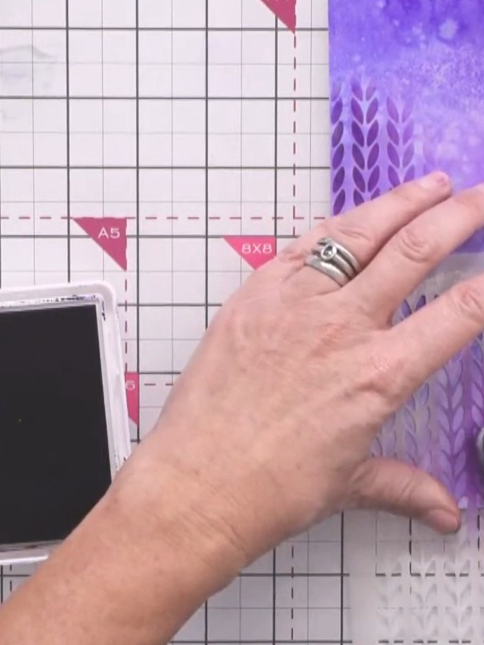 Step 8: Position the Nordic knitted pattern stencil over the purple inky background. Using a brush, apply indigo ink t