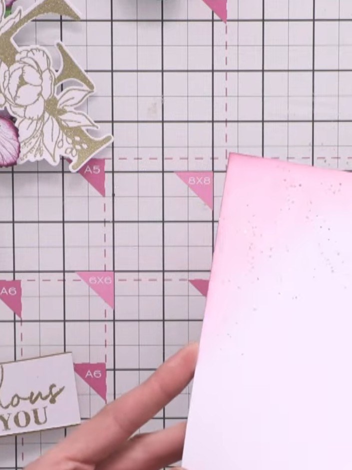 Step 10: Position and adhere the embossed gold F panel onto the pink speckled card base, placing it towards the upper p