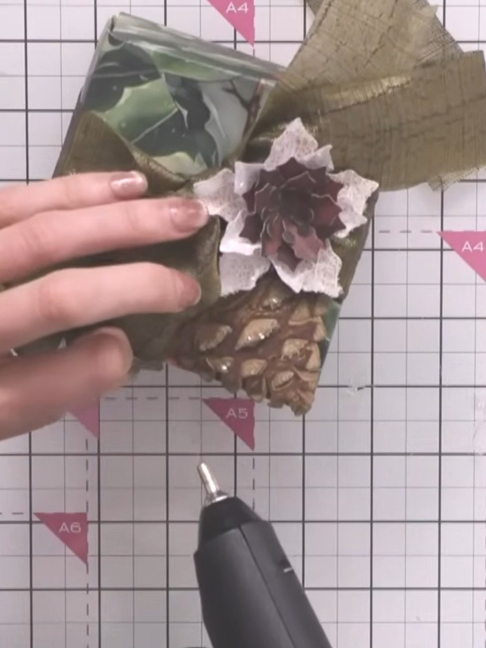Step 7: Arrange all the decorative elements on top of the box, positioning the pine cone, flowers, ribbon bow and foli