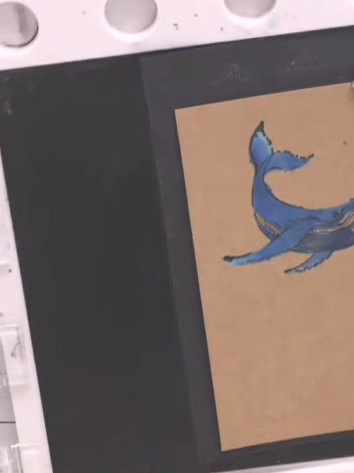 Step 9: Stamp the blue whale image over the white whale base on the kraft card, positioning it to create a layered eff