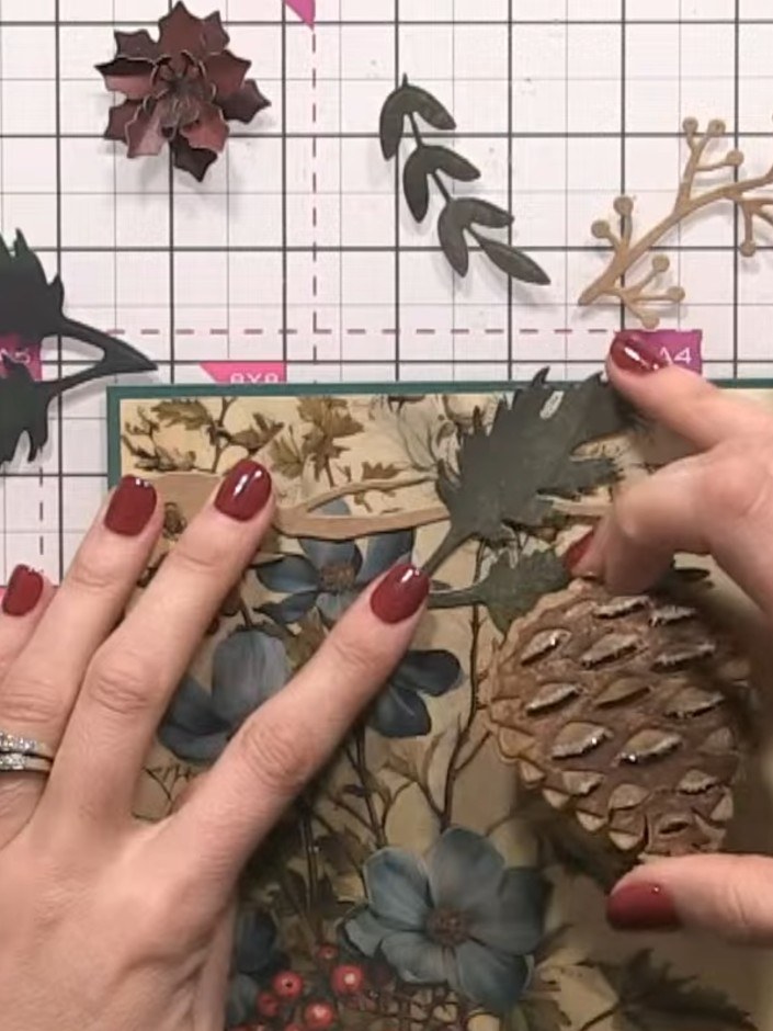 Step 9: Layer the dark green and burgundy poinsettia flowers around the pine cone on the card front. Arrange them in a