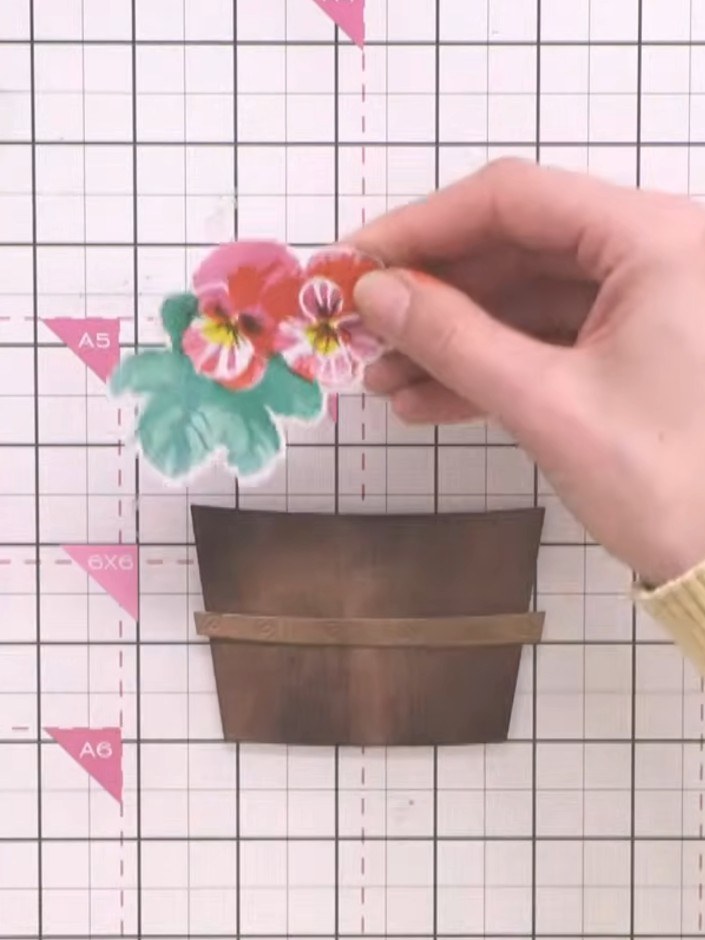 Step 9: Position the die-cut pansy flower clusters above the wooden crate, layering them with 3D foam pads at varying 