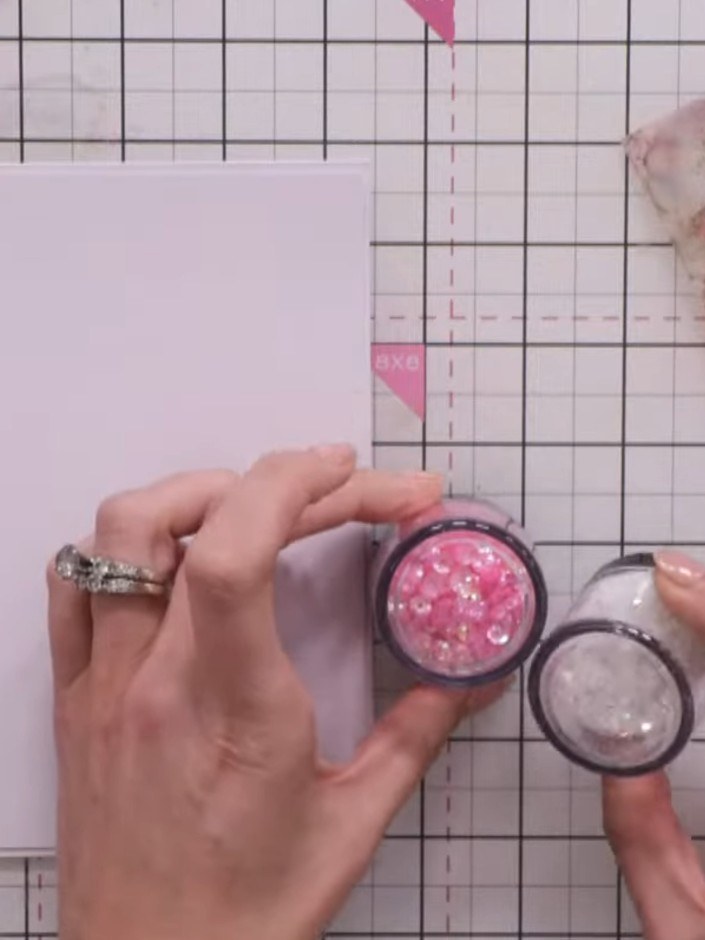 Step 8: Add your shaker contents onto the paper backing inside the aperture. Use pink and white sequins and chunky gli