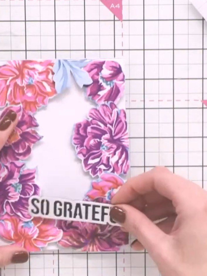 Step 12: Adhere the raised flowers permanently to the card using glue. Add the 'SO GRATEFUL' sentiment die-cut to the c