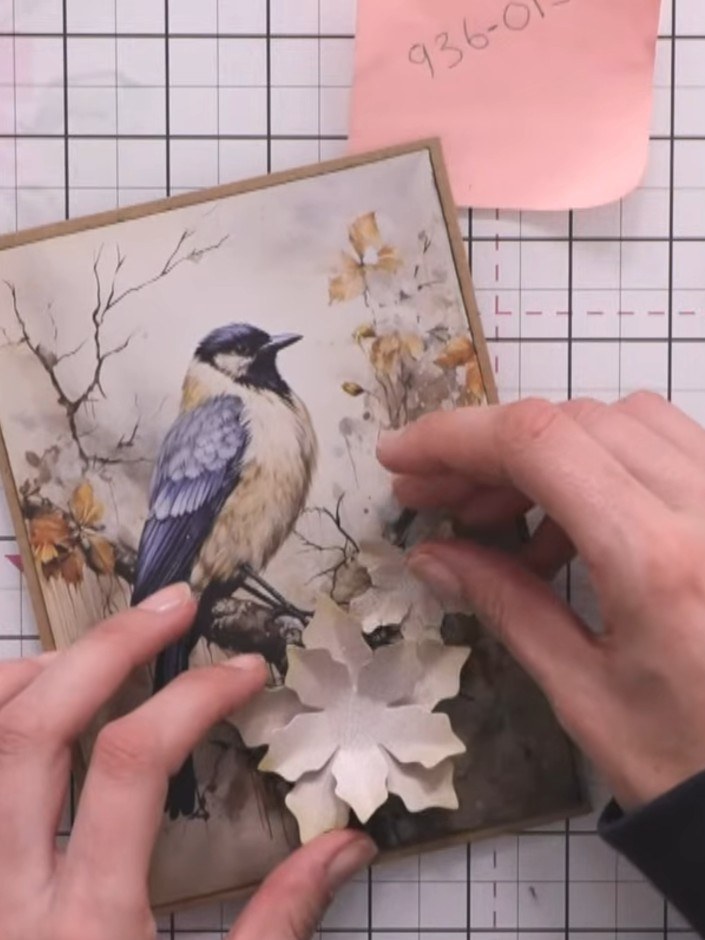 Step 9: Arrange the layered white flowers and dark green foliage on your card with the bird topper, positioning them t