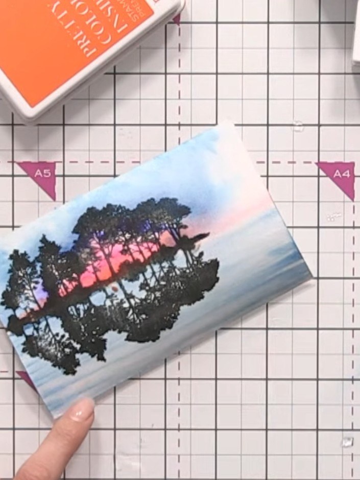 Step 12: Add a subtle touch of orange and pink ink to the water area at the bottom of the card to reflect the sky colou