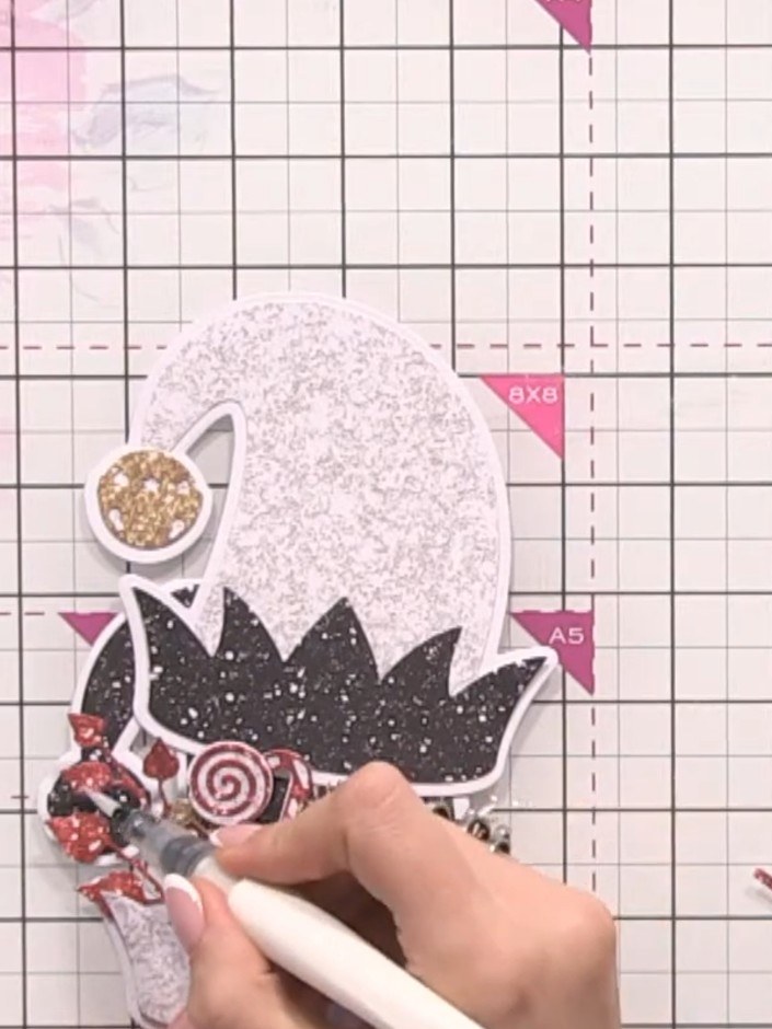 Step 11: Add a small bell embellishment with its band to one of the glittery hats for extra detail. Position the bell t
