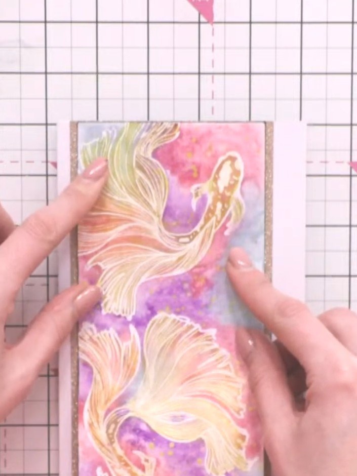 Step 6: Adhere the decorative fish panel with the pink and purple watercolour design onto the gold glitter card base u