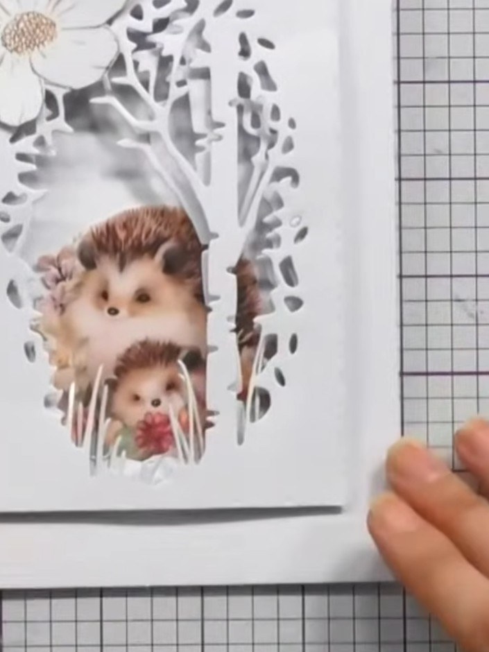 Step 7: Position the hedgehog embellishments within the aperture frame to create a charming scene. Add a white flower 