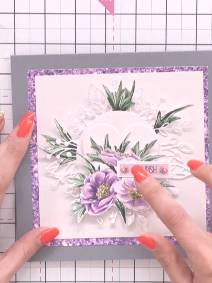 Step 9: Attach a sentiment sticker reading 'Flourish' to the centre of the card, positioning it above the purple flowe