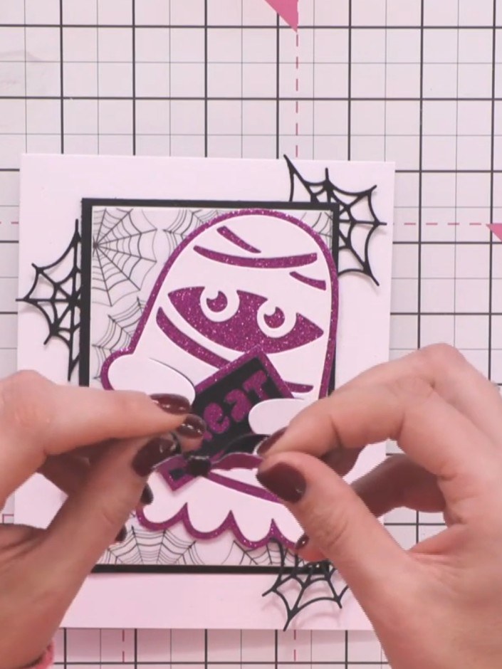 Step 8: Attach the 'TREAT' sentiment piece to the mummy ghost's hands using foam pads or glue. Position it centrally a