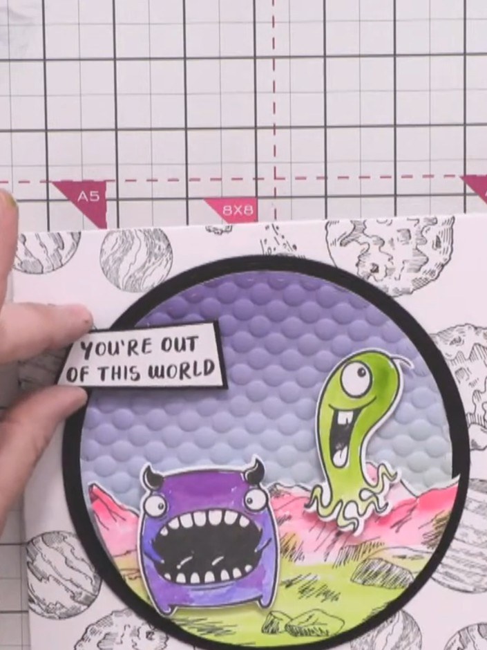Step 12: Adhere the sentiment piece reading "YOU'RE OUT OF THIS WORLD" to the upper portion of the circular card panel 